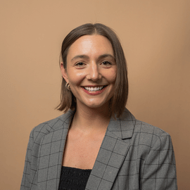 Headshot of a woman smiling at the camera, brown backdrop, gray blazer jacket on